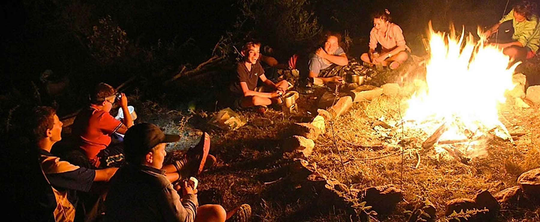 Group of individuals sitting around a campfire at night, engaged in conversation and enjoying the warmth of the flames.