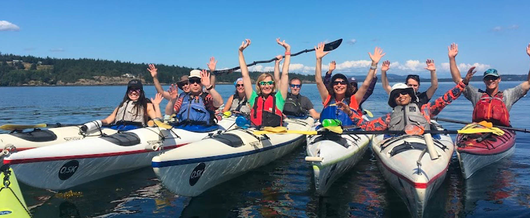 Group of kayakers on the water, smiling and raising their hands while sitting in colorful kayaks during a sunny day.