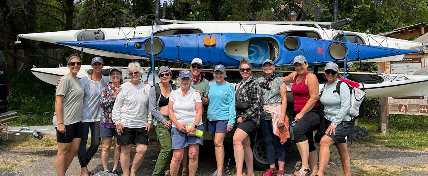 Group of women posing together in front of stacked kayaks, wearing casual outdoor clothing and smiling at the camera.