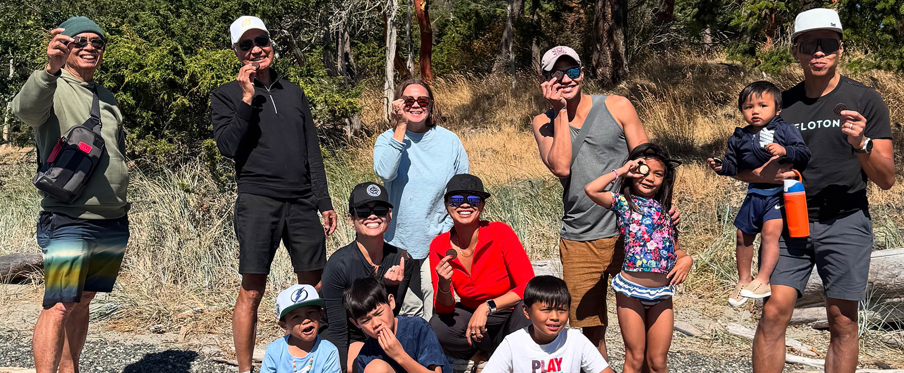 Group of people posing outdoors, some wearing sunglasses and casual clothing, with a mix of adults and children in a natural setting.