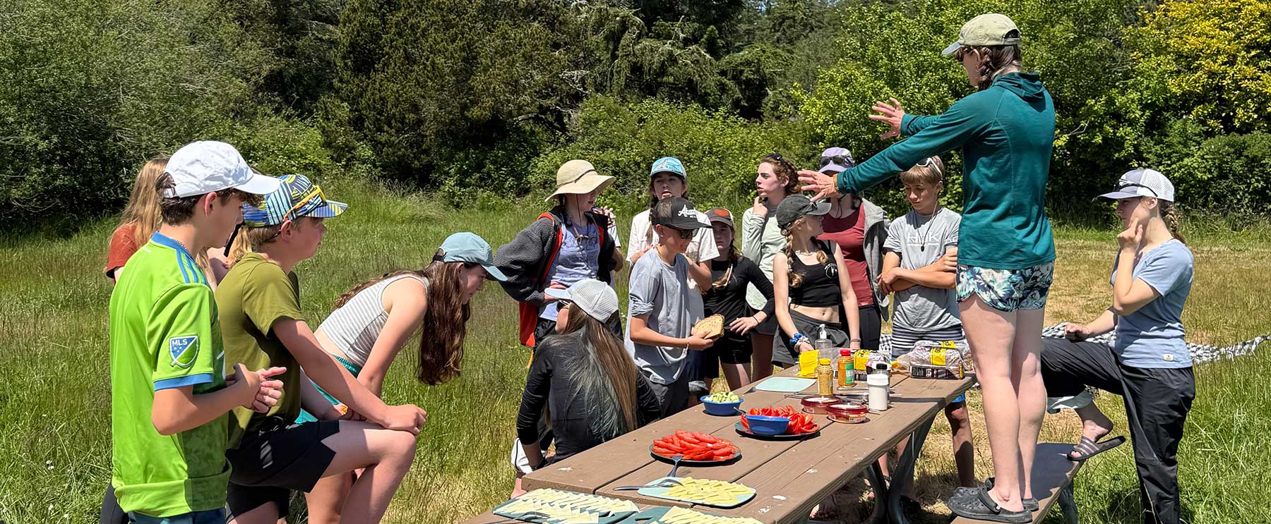 Group tour picnic with Outdoor Odysseys—families in caps gather around a wooden table outdoors, enjoying snacks on San Juan Island grass in summer sunshine.